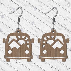 May include: A pair of wooden earrings shaped like a vintage camper van with a mountain and sun cut-out design. The earrings have silver hooks.