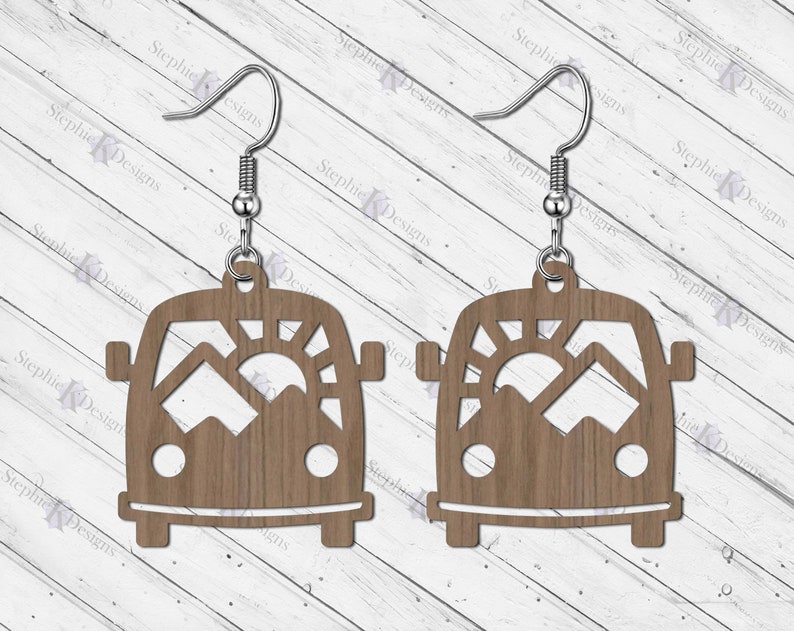May include: A pair of wooden earrings shaped like a vintage camper van with a mountain and sun cut-out design. The earrings have silver hooks.