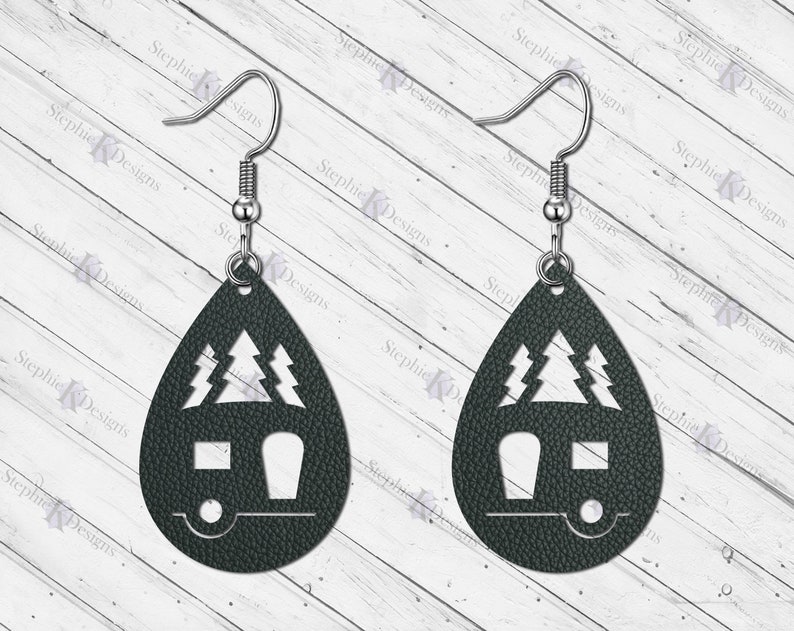 May include: A pair of dark green teardrop-shaped earrings with a camper and pine trees cut out. The earrings are made of leather and have silver hooks.