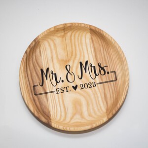 May include: A round wooden tray with the text "Mr. & Mrs. EST. &hearts; 2023" engraved on it.