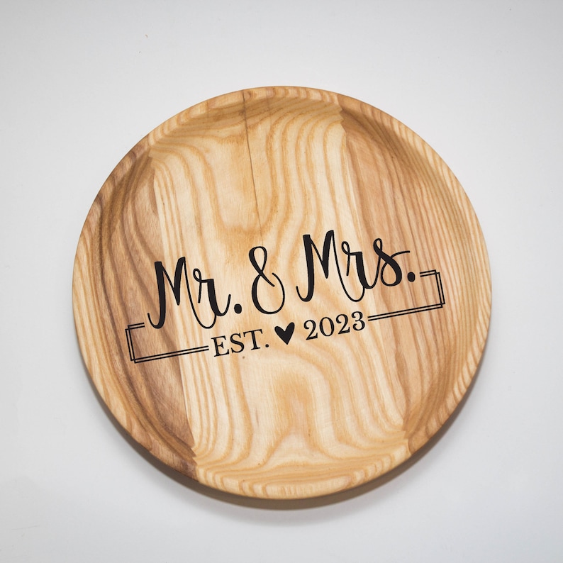 May include: A round wooden tray with the text "Mr. & Mrs. EST. &hearts; 2023" engraved on it.