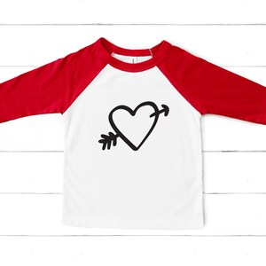 Puede incluir: Una camiseta de b&eacute;isbol blanca y roja con un gr&aacute;fico de coraz&oacute;n y flecha negro.