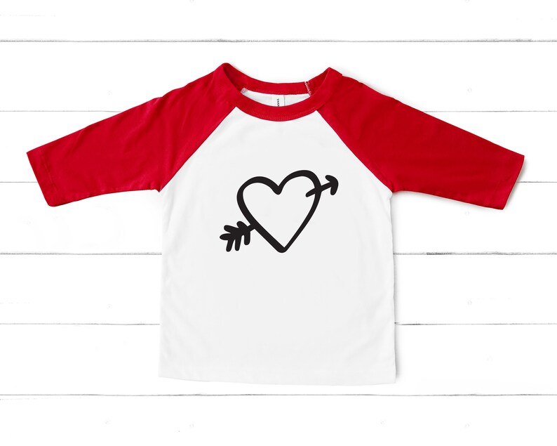 Puede incluir: Una camiseta de b&eacute;isbol blanca y roja con un gr&aacute;fico de coraz&oacute;n y flecha negro.