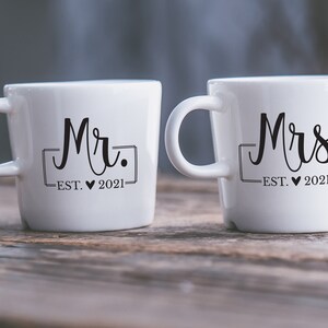 May include: Two white ceramic mugs with black lettering. The mug on the left says "Mr. EST. &hearts; 2021" and the mug on the right says "Mrs. EST. &hearts; 2021".