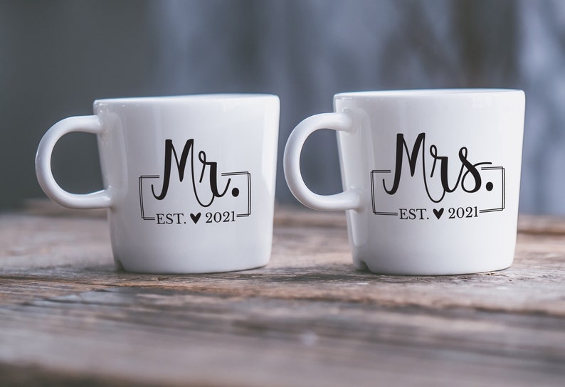 May include: Two white ceramic mugs with black lettering. The mug on the left says "Mr. EST. &hearts; 2021" and the mug on the right says "Mrs. EST. &hearts; 2021".