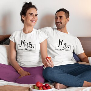 May include: A couple wearing white t-shirts with the text "Mrs. EST. 2021" and "Mr. EST. 2021" printed on them. They are sitting on a bed with white sheets and a gray pillow.
