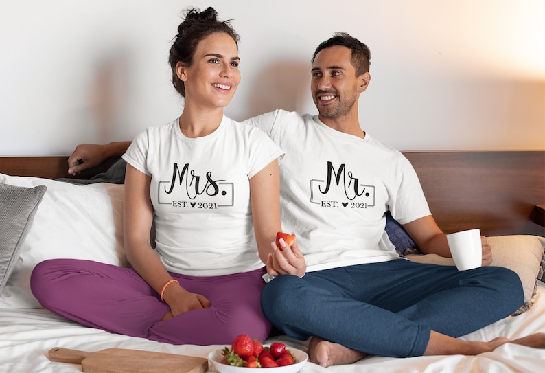 May include: A couple wearing white t-shirts with the text "Mrs. EST. 2021" and "Mr. EST. 2021" printed on them. They are sitting on a bed with white sheets and a gray pillow.