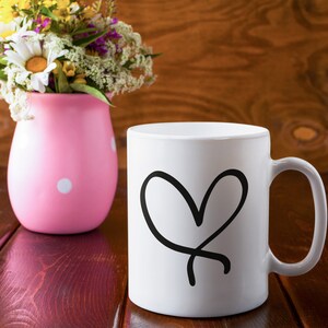 Puede incluir: Una taza de cer&aacute;mica blanca con un dise&ntilde;o de coraz&oacute;n negro dibujado a mano. La taza est&aacute; sobre una mesa de madera junto a un jarr&oacute;n rosa con lunares y flores.