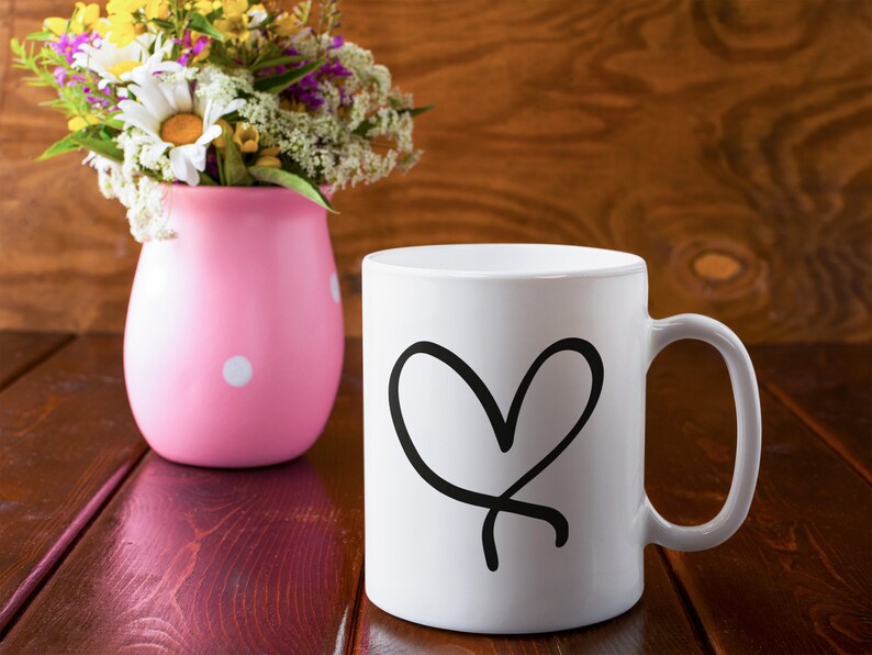 Puede incluir: Una taza de cer&aacute;mica blanca con un dise&ntilde;o de coraz&oacute;n negro dibujado a mano. La taza est&aacute; sobre una mesa de madera junto a un jarr&oacute;n rosa con lunares y flores.