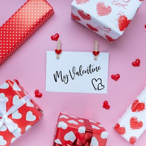 Puede incluir: Un fondo rosa con cuatro cajas de regalo rojas y blancas envueltas en papel con corazones. Una tarjeta blanca con el texto "My Valentine" est&aacute; fijada al fondo con dos pinzas para la ropa. Tambi&eacute;n hay peque&ntilde;os corazones rojos esparcidos alrededor de las cajas y la tarjeta.