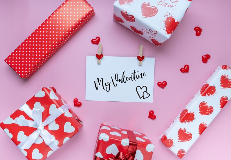 Puede incluir: Un fondo rosa con cuatro cajas de regalo rojas y blancas envueltas en papel con corazones. Una tarjeta blanca con el texto "My Valentine" est&aacute; fijada al fondo con dos pinzas para la ropa. Tambi&eacute;n hay peque&ntilde;os corazones rojos esparcidos alrededor de las cajas y la tarjeta.