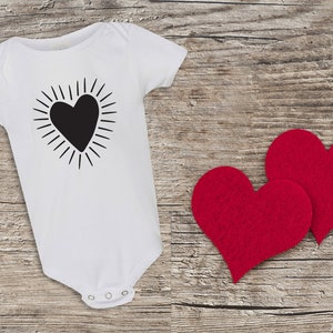 Puede incluir: Un body blanco para beb&eacute; con un gr&aacute;fico de coraz&oacute;n negro con rayos de sol alrededor. Dos corazones de fieltro rojo est&aacute;n sobre un fondo de madera.