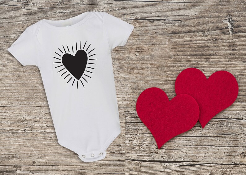 Puede incluir: Un body blanco para beb&eacute; con un gr&aacute;fico de coraz&oacute;n negro con rayos de sol alrededor. Dos corazones de fieltro rojo est&aacute;n sobre un fondo de madera.