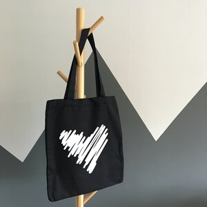 Puede incluir: Una bolsa de tela negra con un dise&ntilde;o de coraz&oacute;n blanco. El coraz&oacute;n est&aacute; hecho de l&iacute;neas garabateadas.