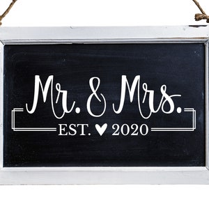 May include: A white framed chalkboard sign with the text "Mr. & Mrs. EST. &hearts; 2020" written in white chalk.