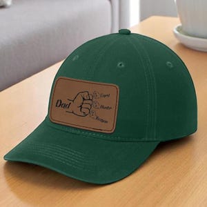 May include: A dark green baseball cap with a curved brim. The cap features a brown leather patch with the word "Dad" and a hand gesture, along with three names. The cap is sitting on a wooden surface.