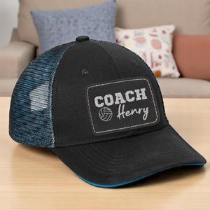 May include: Black and blue trucker hat with the text "COACH Henry" on a patch. The hat features a mesh back and a curved brim. The hat is sitting on a wooden table.