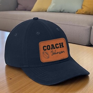 May include: Navy blue baseball cap with a curved brim. A rectangular patch with the words "COACH Johnson" and a football graphic is centered on the front. The cap is sitting on a wooden table.