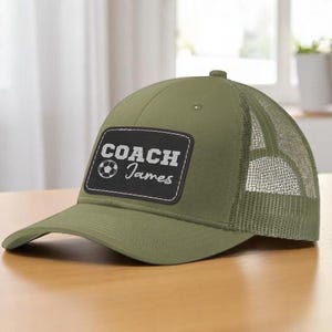 May include: Olive green baseball cap with a mesh back. The front features a black patch with the words "COACH James" and a soccer ball graphic. The cap is sitting on a wooden surface.