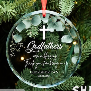 May include: A clear glass Christmas ornament with a white cross and the text "Godfathers are a blessing. Thank you for being mine."  The ornament also includes the name "GEORGE BROWN" and the date "02.24.2024".
