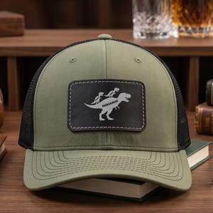 May include: Olive green trucker hat with a black mesh back and a curved brim. The hat features a black leather patch with a white illustration of a dinosaur carrying two figures, and the word "Papasaurus".