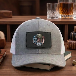 May include: Gray trucker hat with a white mesh back and a curved brim. The front features a black patch with the word "DAD" in red, white, and blue, along with military-themed graphics.