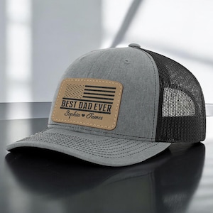 May include: Gray and black trucker hat with a tan leather patch. The patch features a black American flag design and the text "BEST DAD EVER" with the names "Sophia" and "James" below. The hat has a curved brim.