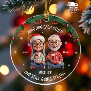 May include: A round clear acrylic Christmas ornament featuring a cartoon illustration of an older couple sitting together. The text reads ANNOYING EACH OTHER, Emma, David, SINCE 1981, AND STILL GOING STRONG. The design includes holly, berries, and snowflakes.