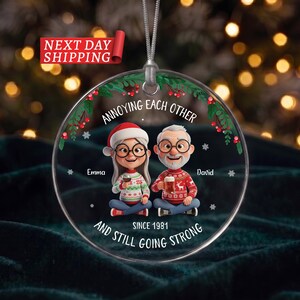 May include: A round clear acrylic Christmas ornament featuring a cartoon illustration of a couple with glasses wearing holiday jumpers. The text reads ANNOYING EACH OTHER, Emma, David, and AND STILL GOING STRONG SINCE 1981, with holly accents.