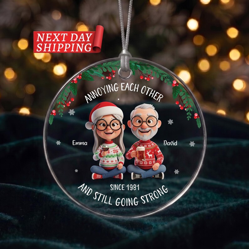 May include: A round clear acrylic Christmas ornament featuring a cartoon illustration of a couple with glasses wearing holiday jumpers. The text reads ANNOYING EACH OTHER, Emma, David, and AND STILL GOING STRONG SINCE 1981, with holly accents.