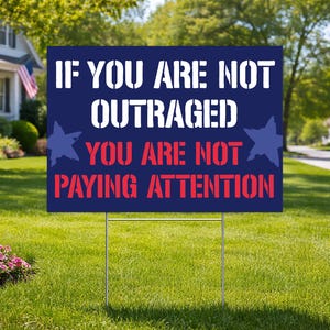 May include: A navy blue yard sign with white text that reads "IF YOU ARE NOT OUTRAGED YOU ARE NOT PAYING ATTENTION." The sign has two blue star graphics and is displayed on a green lawn.