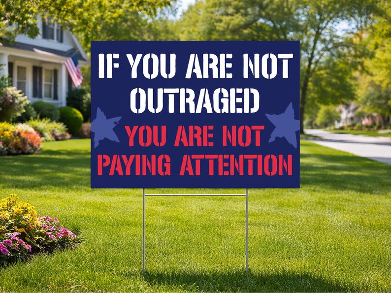May include: A navy blue yard sign with white text that reads "IF YOU ARE NOT OUTRAGED YOU ARE NOT PAYING ATTENTION." The sign has two blue star graphics and is displayed on a green lawn.