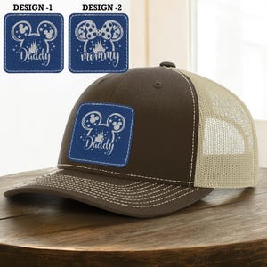 May include: A brown and tan trucker hat with a blue square patch featuring a Mickey Mouse design and the word "Daddy" in white. The hat has a mesh back and a curved brim.