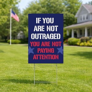 May include: A dark blue yard sign with white and red text that reads "IF YOU ARE NOT OUTRAGED YOU ARE NOT PAYING ATTENTION." The sign is on a metal frame, set in a grassy yard with a house and American flag in the background.
