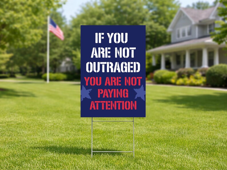 May include: A dark blue yard sign with white and red text that reads "IF YOU ARE NOT OUTRAGED YOU ARE NOT PAYING ATTENTION." The sign is on a metal frame, set in a grassy yard with a house and American flag in the background.