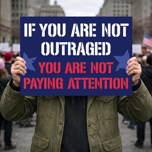 May include: A person holds a navy blue sign with white and red text that reads: "IF YOU ARE NOT OUTRAGED YOU ARE NOT PAYING ATTENTION." The sign has blue star graphics and is held in front of a crowd of people.