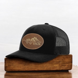 May include: Black trucker hat with a mesh back and a curved brim. The hat features a brown oval patch with a mountain and tree design, and the name "JACK" is printed on the patch.