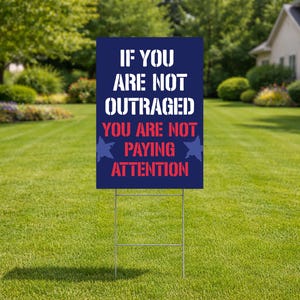 May include: A navy blue yard sign with white text that reads "IF YOU ARE NOT OUTRAGED YOU ARE NOT PAYING ATTENTION." The sign has a metal stake and is placed in a grassy yard.