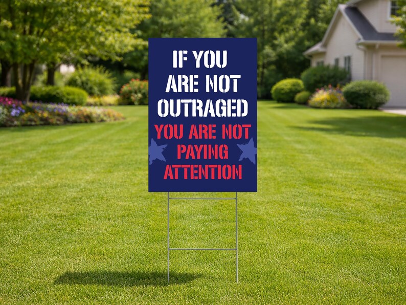 May include: A navy blue yard sign with white text that reads "IF YOU ARE NOT OUTRAGED YOU ARE NOT PAYING ATTENTION." The sign has a metal stake and is placed in a grassy yard.
