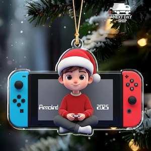 May include: Christmas ornament featuring a cartoon boy wearing a Santa hat, sitting in front of a video game console. The console has blue and red controllers. The name "Arnold" and the year "2025" are displayed on the screen.