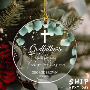 May include: A round, clear glass ornament with a white cross and green eucalyptus leaves. The ornament has the text "Godfathers are a blessing. Thank you for being mine" and "GEORGE BROWN 02.24.2024".