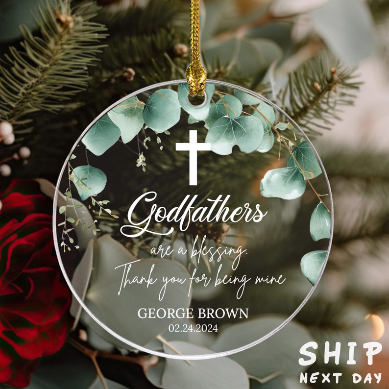 May include: A round, clear glass ornament with a white cross and green eucalyptus leaves. The ornament has the text "Godfathers are a blessing. Thank you for being mine" and "GEORGE BROWN 02.24.2024".
