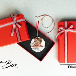 May include: A round, silver ornament with a photo of a family. The ornament has the text "Heaven gained an angel" and a heart. The ornament is in a red gift box with a silver ribbon. The gift box is 120 mm wide and 200 mm tall.