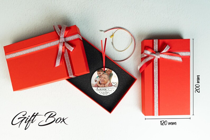 May include: A round, silver ornament with a photo of a family. The ornament has the text "Heaven gained an angel" and a heart. The ornament is in a red gift box with a silver ribbon. The gift box is 120 mm wide and 200 mm tall.