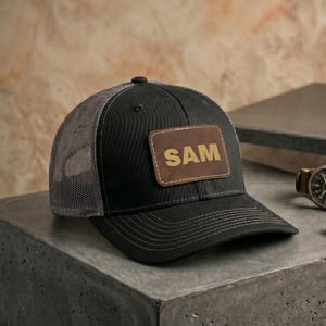 May include: Black trucker hat with a gray mesh back and a brown leather patch. The patch features the name "SAM" in gold lettering. The hat has a curved brim and a snap closure.
