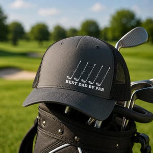 May include: A charcoal gray trucker hat with a black mesh back, featuring white golf club graphics and the text "BEST DAD BY PAR." The hat is resting on a golf bag filled with clubs, set against a green golf course.