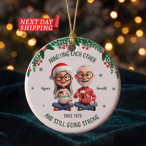 May include: A round white ceramic Christmas ornament featuring a cartoon illustration of an older couple, Agnes and Arnold, wearing holiday jumpers. The text reads: ANNOYING EACH OTHER SINCE 1979 AND STILL GOING STRONG. It has a gold hanging cord.