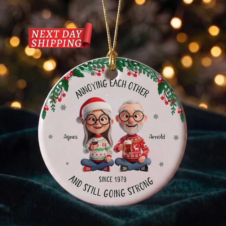 May include: A round white ceramic Christmas ornament featuring a cartoon illustration of an older couple, Agnes and Arnold, wearing holiday jumpers. The text reads: ANNOYING EACH OTHER SINCE 1979 AND STILL GOING STRONG. It has a gold hanging cord.