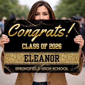 May include: A black graduation sign with gold glitter accents and text. The sign reads "Congrats!" in gold script, "CLASS OF 2026", "ELEANOR", and "SPRINGFIELD HIGH SCHOOL".