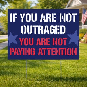 May include: A dark blue yard sign with white and red text that reads "IF YOU ARE NOT OUTRAGED YOU ARE NOT PAYING ATTENTION." Two blue stars flank the text. The sign is displayed on a green lawn.